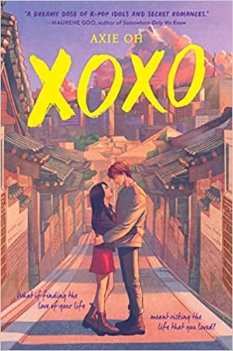 XOXO by Axie Oh book cover features a young couple standing centre‐frame, the girl looking up at the boy as they embrace under a softly glowing sky. The setting behind them is a stylised alleyway of what appears to be Seoul’s traditional architecture—roof eaves and brick paths suggested through gentle warm tones. Overhead, the title “XOXO” is large and bold in vibrant yellow brush‑stroke lettering, instantly catching the eye and conveying both affection (hugs and kisses) and youthful energy.