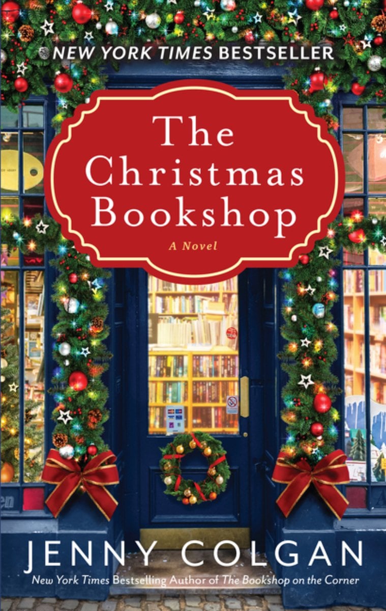 The Christmas Bookshop by Jenny Colgan book cover featuring a charming blue bookshop storefront adorned with festive garland, twinkling lights, red ornaments, and holiday wreaths with velvet bows. The cozy romance novel's cover depicts warm glowing windows revealing bookshelves inside, evoking the magical atmosphere of European Christmas markets and Edinburgh's cobblestone streets.