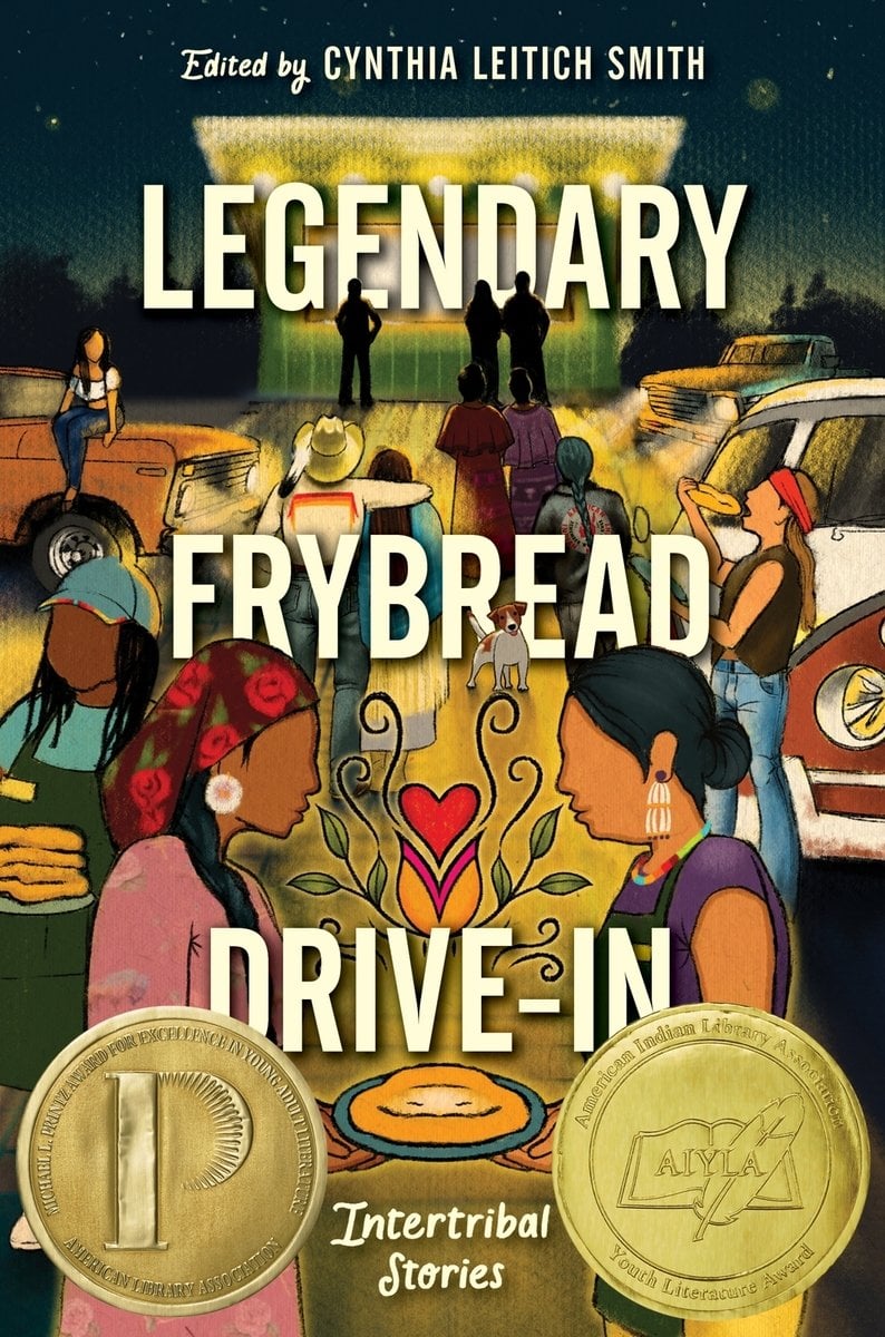 The book cover for Legendary Frybread Drive-In, edited by Cynthia Leitich Smith. At night time, a walk up counter with a green facade and bright lights is set in front of a road lit yellow by headlights of parked cars. Multiple Indigenous figures walk towards the counter, lean on cars, or eat frybread. In the four ground are two women holding a plate of frybread. The woman on the left has brown skin, braided black hair, a burgundy hair scarf with a rose pattern, a pink floral dress, and white daisy earrings. The woman on the right has olive skin, black hair pulled into a bun, a purple top, a yellow skirt, and a green work apron. Flowering between them is a yellow and red heart with a red heart nestled between its petals.