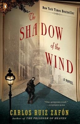 Book cover of spanish author. Title "The Shadow of the Wind" is written in red. The cover looks like it is a page folded in the center. At the bottom is a man looking over his shoulder and a streetlamp.