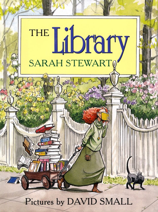 The Library by Sarah Stewart, Illustrated by David Small, book cover. A young woman with red curly hair and wearing a green dress has her face covered by a book while she is pushing a wagon filled with books, she walks down a sidewalk in her town. The background is a white picket fence and flowery bushes.