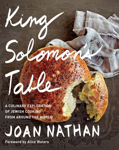 Book jacket for King Solomon's Table: A Culinary Exploration of Jewish Cooking from Around the World: A Cookbook by Joan Nathan. On a brown table overlaid with a transparent map, a lighter brown cloth lays next to a circular loaf of torn bread.
