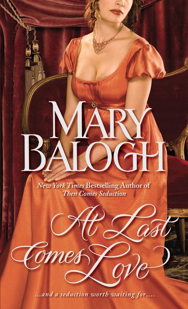 At Last Comes Love by Mary Balogh, a historical romance book cover depicting a woman wearing an orange, short-sleeved Regency era gown, sitting in a maroon colored chair. The title is printed in a bold, cursive, white font.