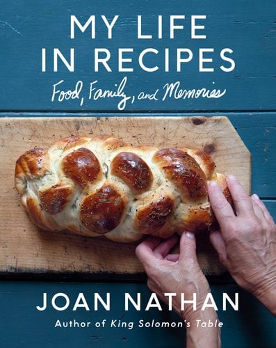 Book jacket for My Life in Recipes: Food, Family, and Memories by Joan Nathan. On a blue wooden table, a wooden cutting board holds a braided loaf of bread. Two hands reach to tear a piece away.