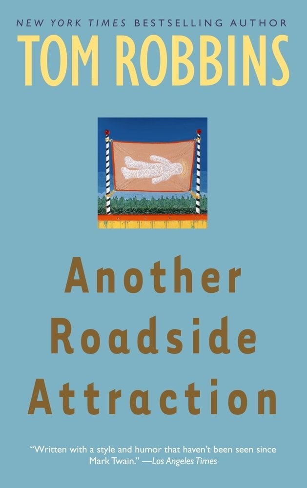 Another Roadside Attraction by Tom Robbins book cover depicting a light blue background with a small image in the center of the top half. The image is of a banner with a mummy on it between two striped poles standing in a grassy field.