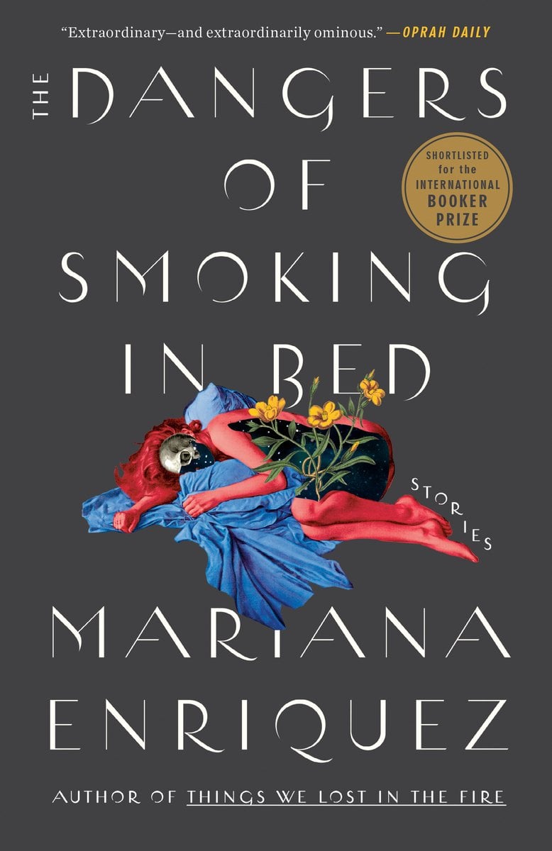 Cover of The Dangers of Smoking in Bed by Mariana Enriquez. Grey background with a red skeleton that has skin on some areas, laying on the ground. The person is hugging a blue shirt and has flowers growing out of her.