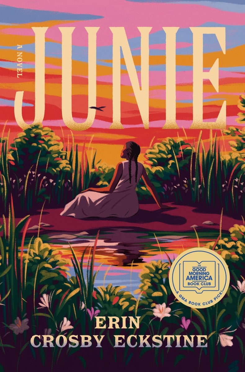 Junie by Erin Crosby Eckstine. A young girl sits on a grassy riverbank at sunset, surrounded by tall reeds and flowers. The sky glows in pink, orange, and purple tones, reflected in the calm water