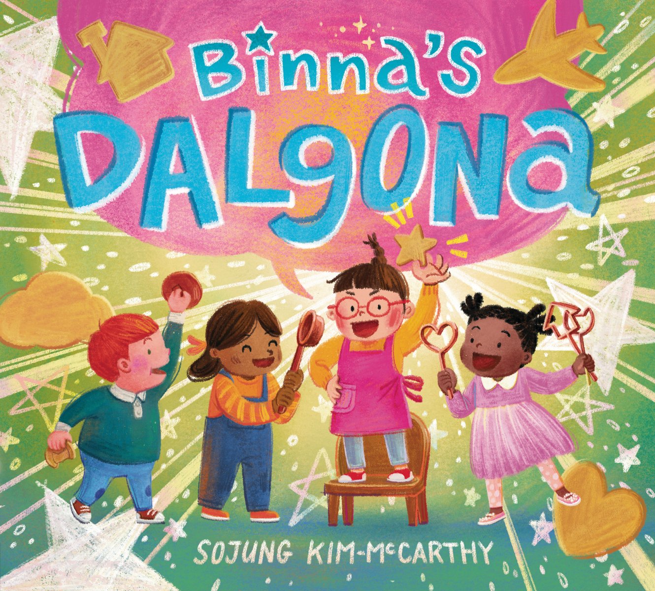Binna's Dalgona by Sojung Kim-McCarthy, book cover. A group of four young children—three girls and one boy—stand together holding cookie cutters, bowls, and spoons. One of the girls in a pink apron and yellow shirt stands on a brown chair with her arm raised, holding a star. A giant pink speech bubble connects to her with the title largely in blue.