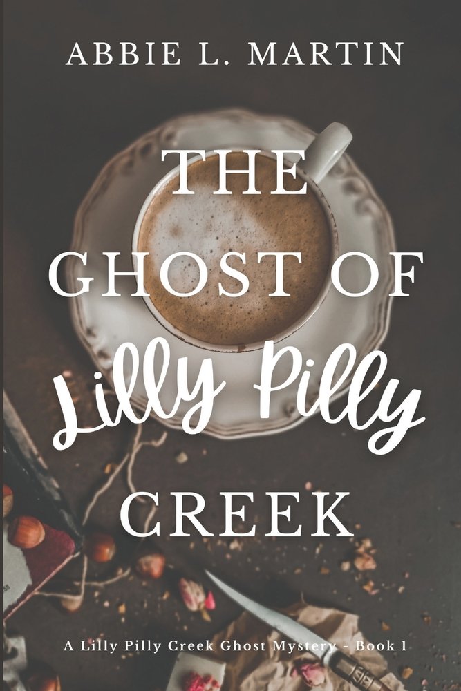 The Ghose of Lilly PIlly Creek by Abbie L. Martin cozy fantasy book cover depicting a cup of cappuccino on a lacy white plate set on a butcher's block with assorted crumbs, a knife, and berries.