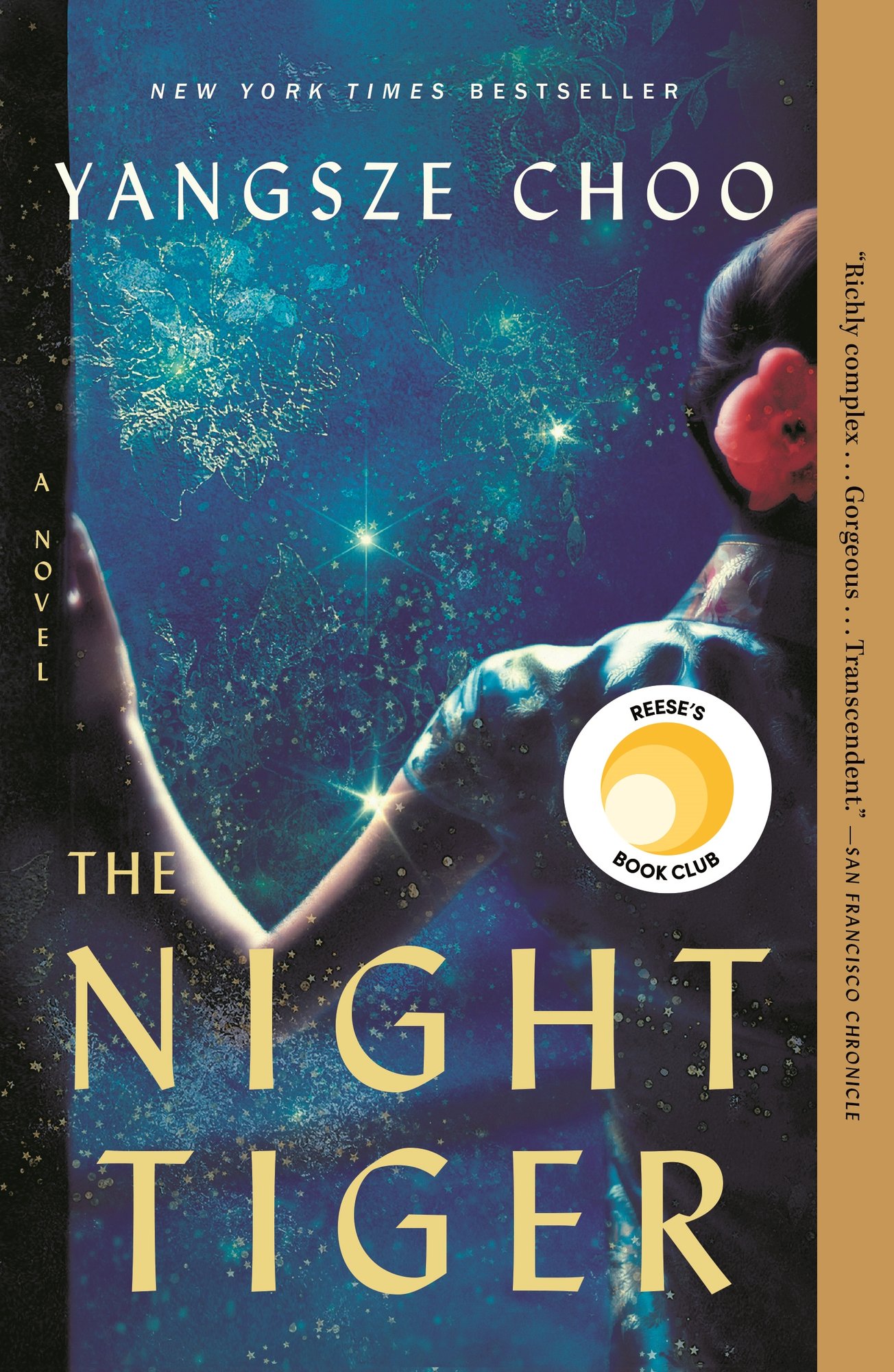 The Night Tiger by Yangsze Choo, sparkly blue book cover featuring a woman with dark hair and a pink flower in it opening a door.