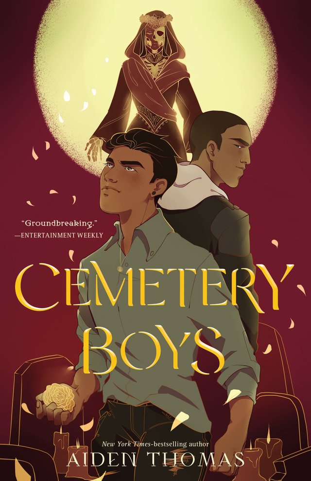 Cemetery Boys by Aiden Thomas. Book cover. Artwork of two Latinx boys posing back-to-back while one of the boys holds a golden flower. A skeleton woman (Lady De La Muerta) looms above them in front of a full yellow moon. There are gravestones and candles at the bottom and golden petals are falling around the boys.
