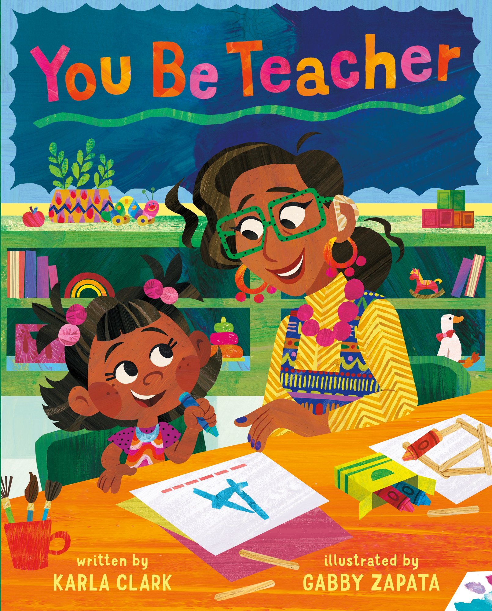 You Be Teacher by Karla Clark, Illustrated by Gabby Zapata, book cover. In a classroom setting with green shelves and a chalkboard in the background, a teacher in colorful clothing and green glasses and dark hair sits with a young girl at a table. The little girl is wearing parts of her hair in pigtails and has a pink shirt on. In front of them, they are writing the letter 'A' on a paper.