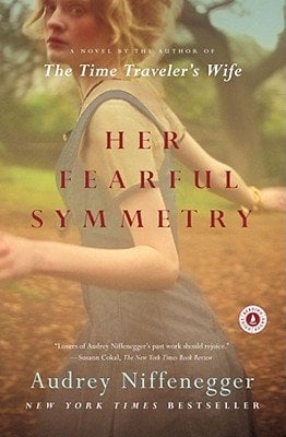Her Fearful Symmetry by Audrey Niffenegger book cover shows a portrait of a young woman from the shoulders up viewed from behind and slightly to the side, her blonde hair loose in curls. She wears a muted blue-gray sleeveless dress and stands in a blurred woodland or cemetery-like setting where autumnal trees with golden-green foliage float in soft focus. The title “Her Fearful Symmetry” appears in large, elegant red serif letters across the center, with Audrey Niffenegger and “A Novel by the Author of The Time Traveler’s Wife” in smaller white type above and below, respectively.
