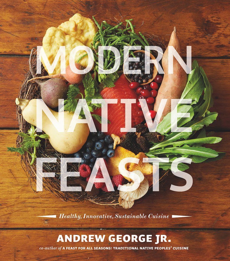 Modern Native Feasts, a cookbook written by the Indigenous chef Andrew George Jr. highlighting Indigenous recipes, book cover of a nest holding natural ingredients like mushrooms, squash, tomatoes, blueberries, leafy greens, raspberries, sweet potato, and salmon on a wooden table with white low opacity title text over the foods.