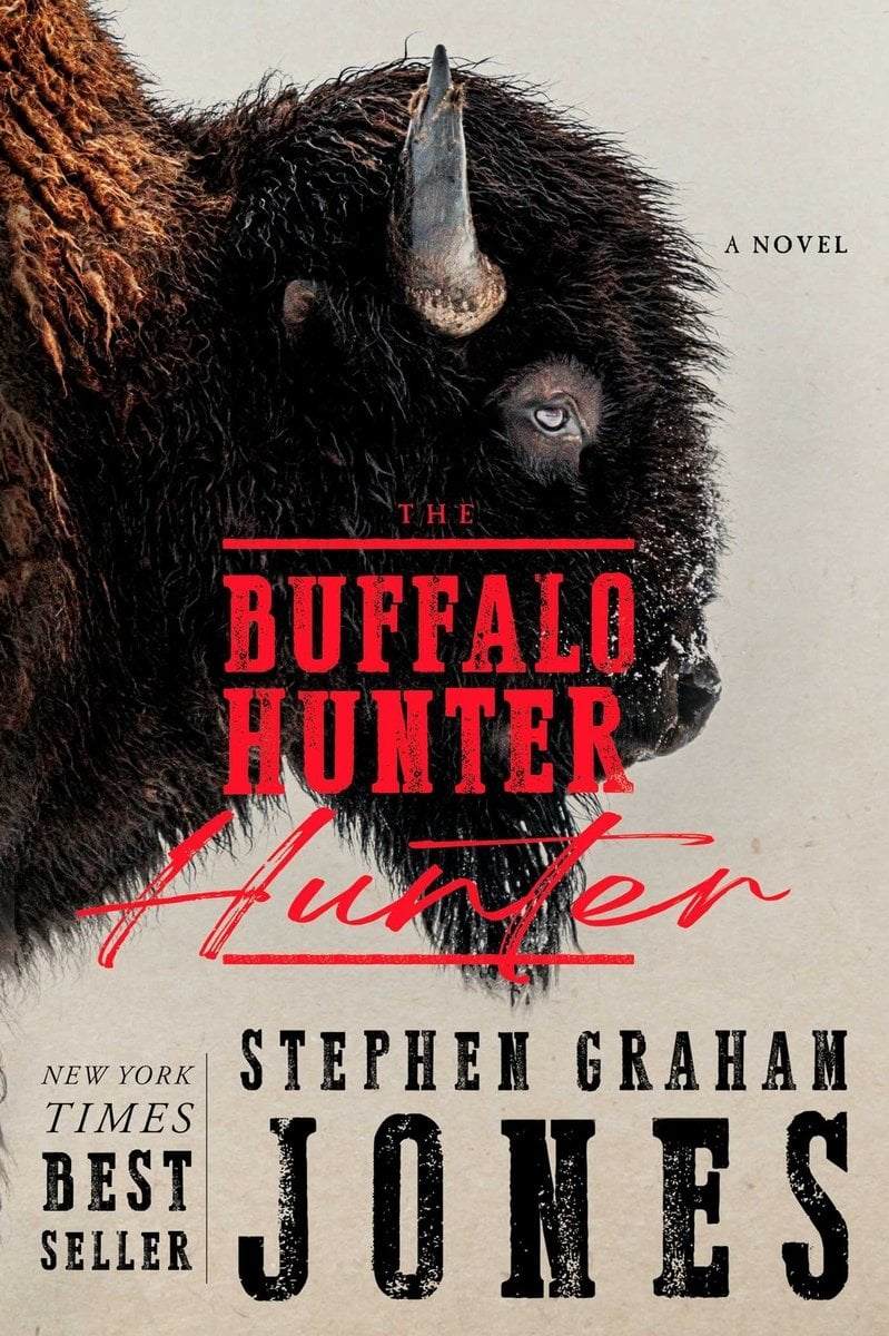 Book cover for "The Buffalo Hunter Hunter" by Stephen Graham Jones. A close-up profile of a buffalo's head dominates the image, showing dark shaggy fur, a curved horn, and one piercing blue eye against a beige background. The title appears in bold red text with "Hunter" deliberately repeated twice. "NEW YORK TIMES BESTSELLER" appears on the left side, "A NOVEL" in the upper right corner, and the author's name in large black letters at the bottom.